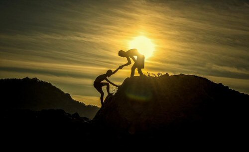 A silhouette image of one adolescent extending a hand to help another adolescent climb a mountain.