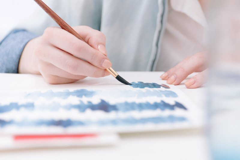 Arts therapy image of a close up of a person's hand using a paintbrush on a white page in an arts therapy context. 